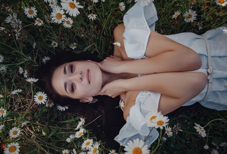Beautiful young brunette woman in a white dress is enjoying spring in a field of daisies. nice female lying down in meadow of flowers, happy young lady and spring green nature, harmony conceptの写真素材
