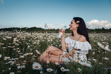 Beautiful young brunette woman in a white dress is enjoying spring in a field of daisies. woman drinking water from a bottle. Health idea and concept, beautiful healthy hair and skin, lack of allergiesの写真素材