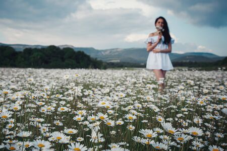 Beautiful young brunette woman in a white dress is enjoying spring in a field of daisies. Health idea and concept, beautiful healthy hair and skin, lack of allergiesの写真素材