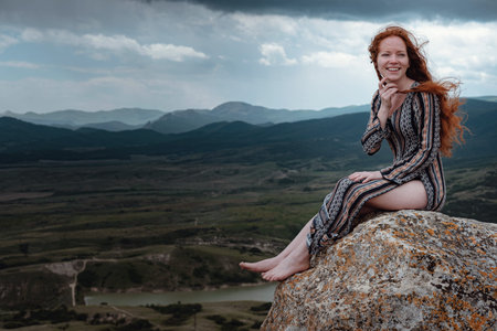 Ginger woman sits on a cliff and looks at an impending storm, watching the last light. Hair blowing...