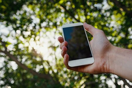 male hand hold white smart phone, cell phone, mobile over blurred image of green forest background.の写真素材