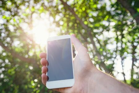 male hand hold white smart phone, cell phone, mobile over blurred image of green forest background.の写真素材