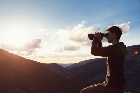 Young man at mountain looking through binoculars with canyon view. Banner with copy space. idea and concept of travel and discoveries, adventures and freedomの写真素材