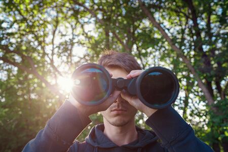 Explorer hiker young man in plaid shirt looking through binoculars on background of summer forest and sea bay outdoor, front view. Travel concept.の写真素材