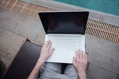travel blogger sitting at swiming pool writing article on white laptop. Freelance remote work concept. Self employed man coding. Copy space, pool view backgroundの写真素材