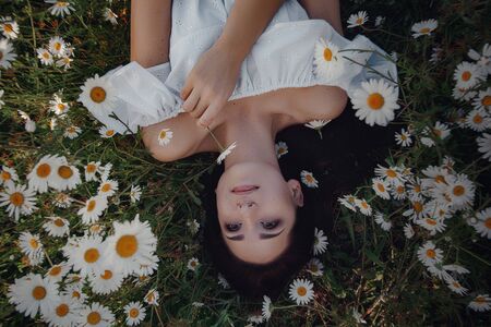 Beautiful young brunette woman in a white dress is enjoying spring in a field of daisies. nice female lying down in meadow of flowers, happy young lady and spring green nature, harmony conceptの写真素材