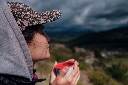 tourist girl in a cap holds a mug with a hot drink in her hands, enjoying the scenery. Beautiful green valley and dramatic rain skyの写真素材
