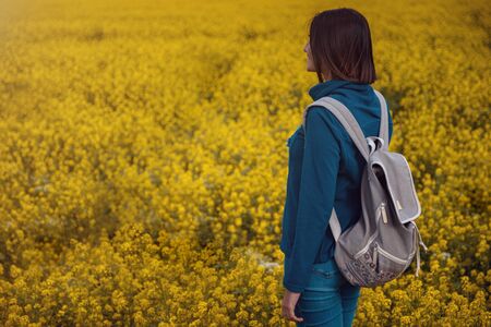 Woman traveler in a blue sweater and jeans in a flowering rapeseed field. Happy beautiful young woman enjoys smell of blooming rapeseed field full of joy and happiness. Concept of freedomの写真素材