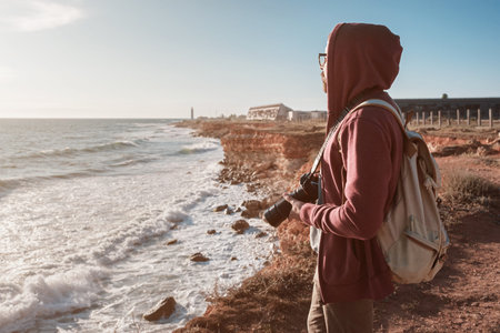 Happy hipster man with backpack standing on the rock and looking at the seashore and beacon at...