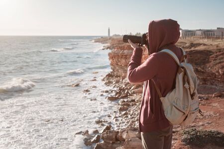 Happy hipster man with backpack standing on the rock and takes a picture of the sea with a camera. Hiking in cold season. Wanderlust concept scene.の写真素材