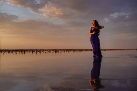 Elegant woman in silky purple dress walking by a salt lake. Romantic mood. Water reflection of clouds and empty space. Holiday, vacation travel scene. glorious sky at sunsetの写真素材