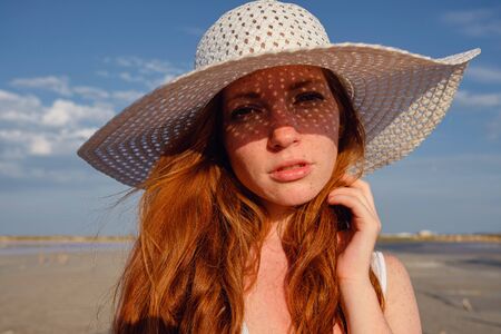 Beautiful young lady with long healthy red hair and cute white dress walking near lake. Hot summer evening. Sunset. Lifestyle concept.の写真素材
