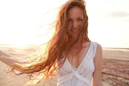 Beautiful young lady with long healthy red hair and cute white dress walking near lake. Hot summer...