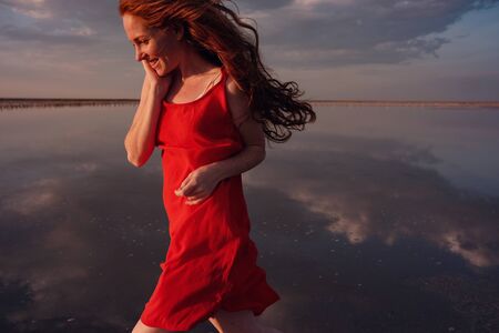 Elegant woman in silky red dress walking by a salt lake. Romantic mood. glorious sky at sunset. lake Sivashの写真素材