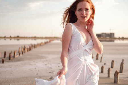 Beautiful young lady with long healthy red hair and cute white dress walking near lake. Hot summer...
