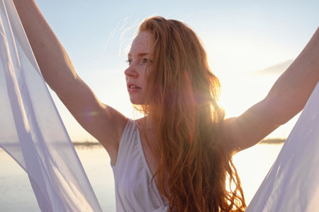 Beautiful young lady with long healthy red hair and cute white dress walking near lake. Hot summer...