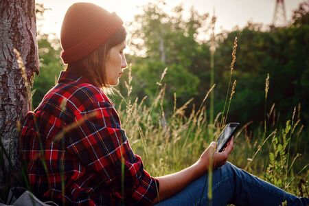 Beautiful female traveler sitting near tree and typing on mobile phone. Trendy young woman in fall in park texting.の写真素材