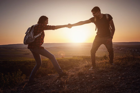 Silhouette in mountains, sunrise. Teamwork of man and woman hiker helping each other on top of...