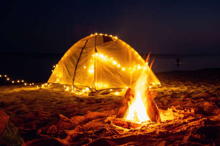 The fire at night on the beach. Summer mood. Night camping with a retro garland on sea shore.の写真素材