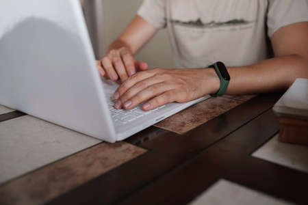Focused young man wearing glasses using laptop, typing on keyboard, writing email or message, chatting, shopping, successful freelancer working online on computer, sitting in kitchen, closeup on handの写真素材