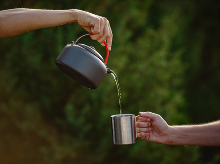 Hiker pouring a hot drink from a metal teapot into a steel mug. Atmospheric tranquil moment.