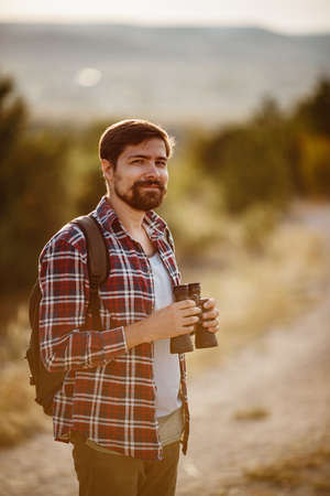 Guy looking at binoculars in hill. man in t-shirt with backpack. Young Caucasian man during hike in valley landscapeの写真素材