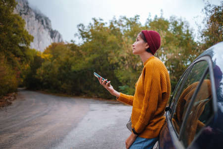 Woman beside a car and looking at smart phone surrounded autumn forest in mountains. Portrait of romantic hipster female, Warm autumn weather, calm scene. Wanderlust photo series.の写真素材