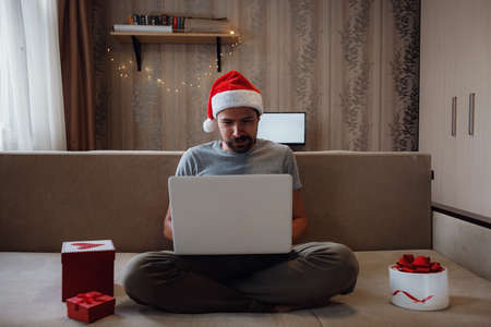A hipster man with a red cup sitting on a sofa at home at Christmas time. Virtual Christmas house party. Online team meeting video conference calling from home.の写真素材