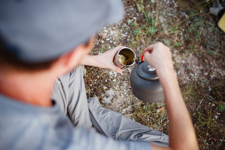 Outdoors image of young explorer man drinking hot beverage in mountains, sitting and relaxing after...