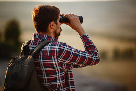 Guy looking at binoculars in hill. man in t-shirt with backpack. Young Caucasian man during hike in valley landscapeの写真素材