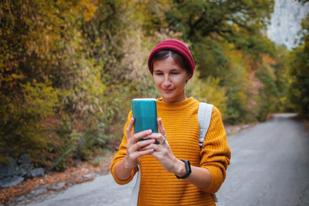 Hipster woman with backpack hold on smart phone gadget in forest. Traveler using mobile on...