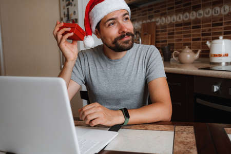 A hipster man with a red cup sitting on a sofa at home at Christmas time. Virtual Christmas house party. Online team meeting video conference calling from home. Man wearing Santa hatの写真素材
