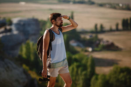 Guy looking at binoculars in hill. man in t-shirt with backpack. Young Caucasian man during hike in valley landscapeの写真素材