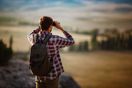 Guy looking at binoculars in hill. man in t-shirt with backpack. Young Caucasian man during hike in valley landscapeの写真素材