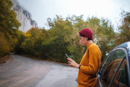 Woman beside a car and looking at smart phone surrounded autumn forest in mountains. Portrait of...