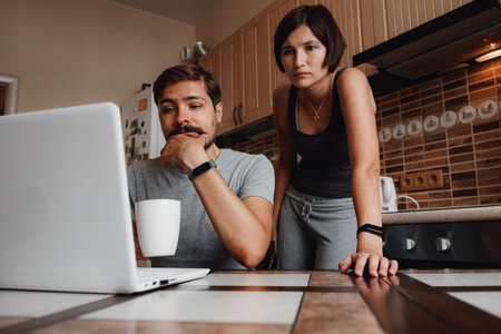 Young couple using laptop and smartphone in the kitchen, smiling man working online at home, family...
