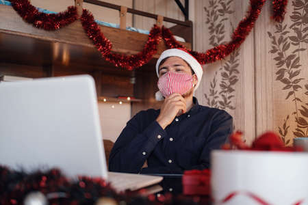 portrait of unhappy man in mask sitting in room near christmas decorations with lights and ordering gifts. New Year e-shopping. Merry Christmas Covid 19 coronavirus social distance concept.の写真素材