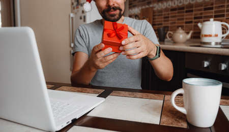 A hipster man with a red cup sitting on a sofa at home at Christmas time. Virtual Christmas house party. Online team meeting video conference calling from home. Man wearing Santa hatの写真素材