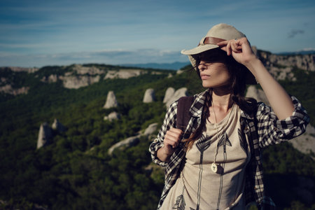 woman traveler with backpack holding hat and looking at amazing mountains and forest, wanderlust travel concept, space for text, atmospheric epic momentの写真素材