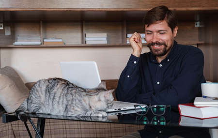Young bearded businessman sits in home office at table and uses laptop, next sits gray cat. On table is smartphone, paper, books, cup of coffee. Working home concept during quarantineの写真素材