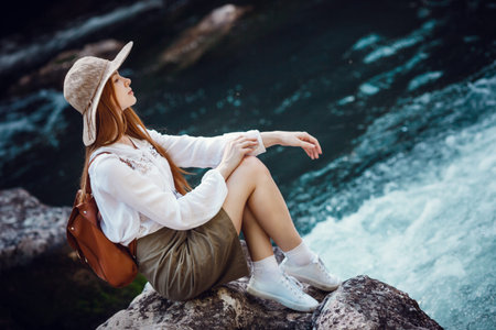 A beautiful young woman with red hair, in a hat and with a backpack near a noisy river in the...