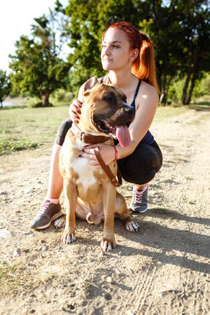 redhair jouful young woman caressing their dog, wearing sport clothing, enjoying their time and vacation in sunny parkの写真素材