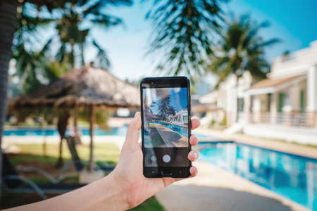 hand holding phone on background of the pool in hotel. photo camera on the screen. close up hand hold smartphone take a picture of summer vacationの写真素材