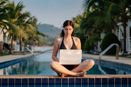 Woman using her laptop on the pool edge on a sunny day. Freelance work in tropical countryの写真素材