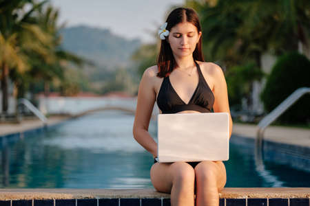 Woman using her laptop on the pool edge on a sunny day. Freelance work in tropical countryの写真素材