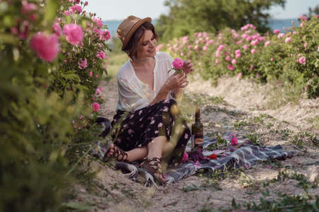 beautiful girl wearing hat with book sitting on grass in rose gaden. A country girl enjoys the sunset, wine and reading books in the fragrant rose garden. The concept of perfumeの写真素材