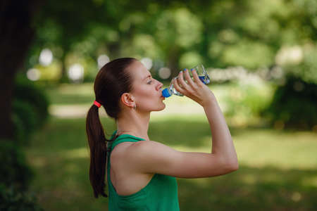 Portrait of young beautiful dark-haired woman wearing green t-shirt drinking water at green park after workout exercising on sunset evening summerの写真素材