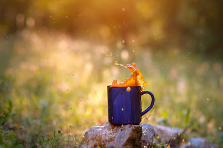 Blue mug of hot tea or coffee with milk, outdoor, the concept of travel, spray, splash. Glare on the pictures from the setting sun, pleasant bokehの写真素材