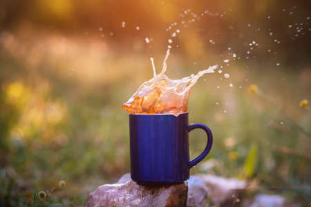 Blue mug of hot tea or coffee with milk, outdoor, the concept of travel, spray, splash. Glare on the pictures from the setting sun, pleasant bokehの写真素材