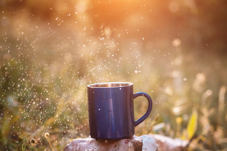 Blue mug of hot tea or coffee with milk, outdoor, the concept of travel, spray, splash. Glare on the pictures from the setting sun, pleasant bokehの写真素材